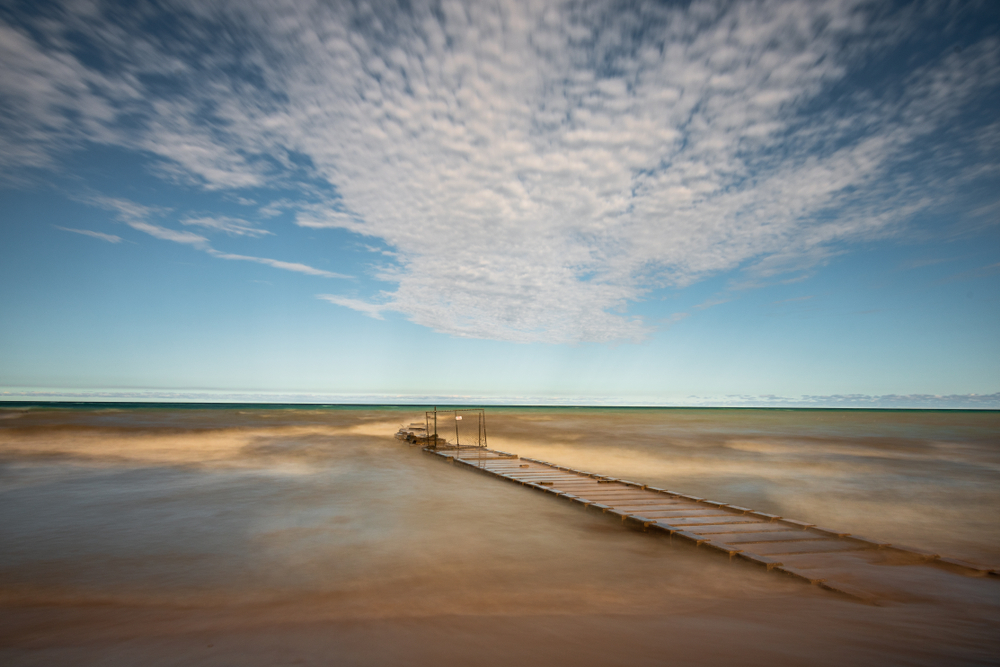 A boardwalk at Atwater Beach. You can see the lake in the distance and some sandy areas around the boardwalk. The sky is very blue with some clouds. 