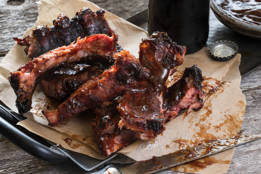 A plate of barbecue ribs. The plate has brown paper on it and there is a knife with barbecue sauce on the plate. You can see a bottle cap, a bottle, and the wooden table top.