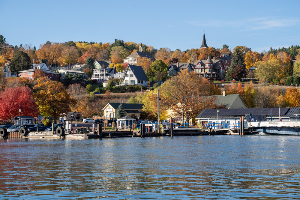 The view of the small town of Bayfield from the lake. It has lots of charming buildings going up the side of the hill and trees with brown, yellow, orange, red, and green leaves. There is also a dock area on the water. 