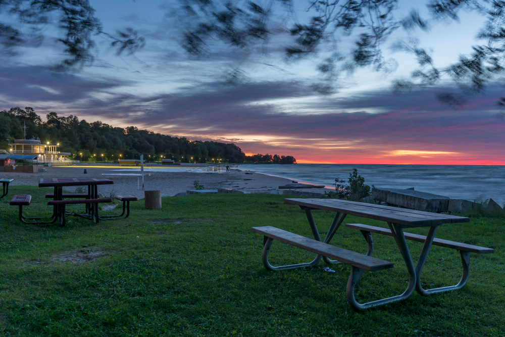 Bradford Beach at twilight. There are picnic tables in a grassy area, a tiki building, and a sandy area. There are a few people on the sandy area. The sky is cloudy but you can see some pink, purple, and orange bits of sky. 