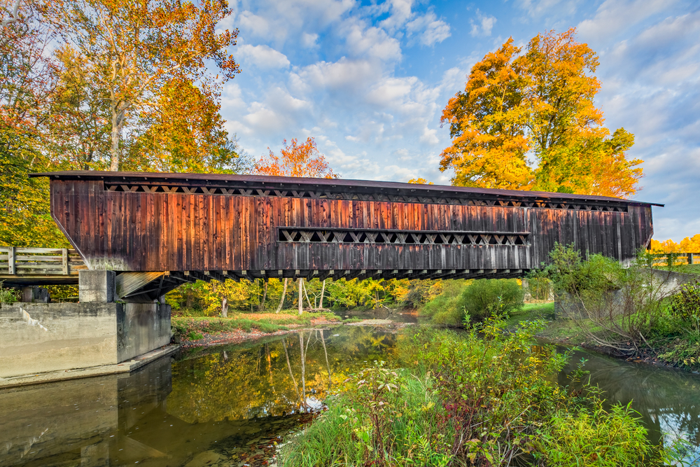 A brown rusty looking covered bridge over the creek with trees in the background