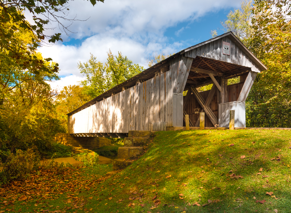 A white covered bridge on a road on a green bank surrounded by trees