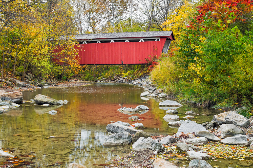 A red bridge over a creek with autumn foliage on either side. One of the covered bridge in Ohio