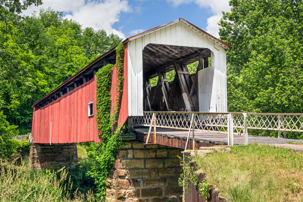 A red covered bridge with a white front and white fence covered in ivy