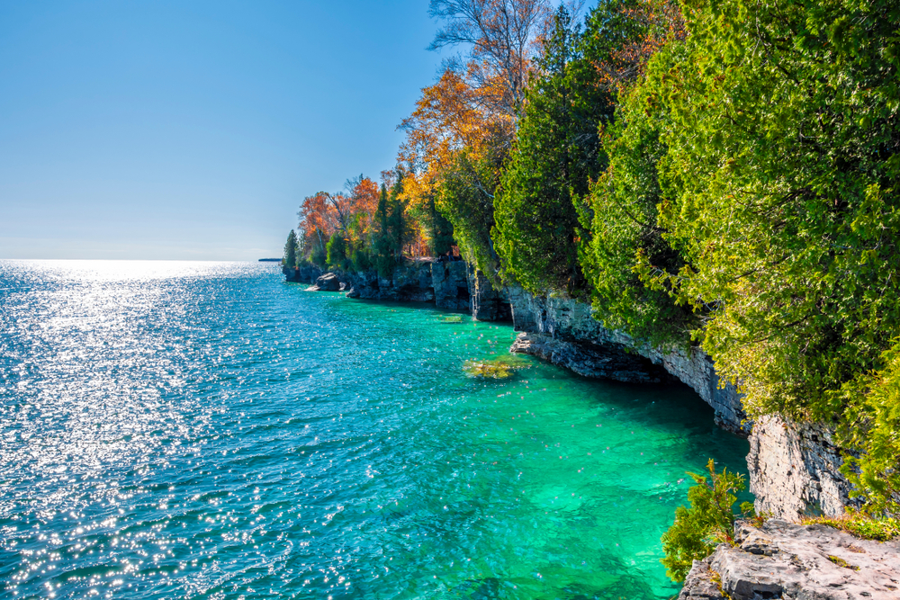 A view looking down the shore of one of the islands at Cave Point State Park. The shore is large rock formations covered in trees with green, yellow, orange, and some red leaves. The water is very blue. 