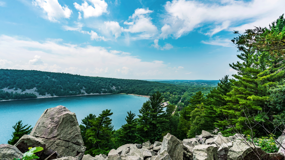 The view looking over a rocky cliff down into a very blue lake in Wisconsin. The lake is surrounded by tall trees with green leaves. The sky is very blue and there are there some clouds. 