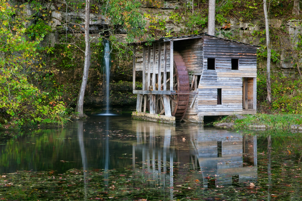 An old wooden slat mill in front of a stone wall. The stone wall has greenery all over it and a small waterfall coming from it. The mill has a rusted wheel and is perched on water. 