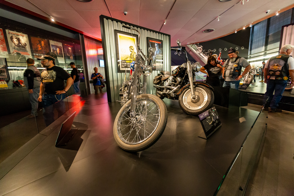 People wearing typical 'biker gear' looking at the motorcycles in the Harley Davidson Museum. There are motorcycles, framed vintage advertisements, and more in the room. 