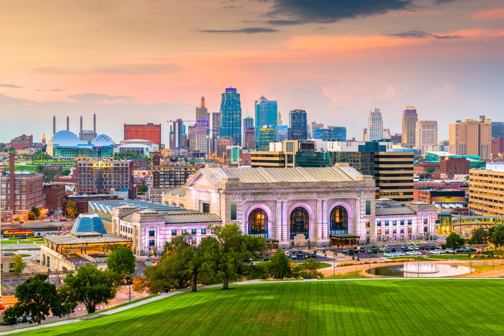 The Kansas City skyline at sunset. The city is lit up but the sky is still very bright. There is a mix of modern and old buildings in the skyline.