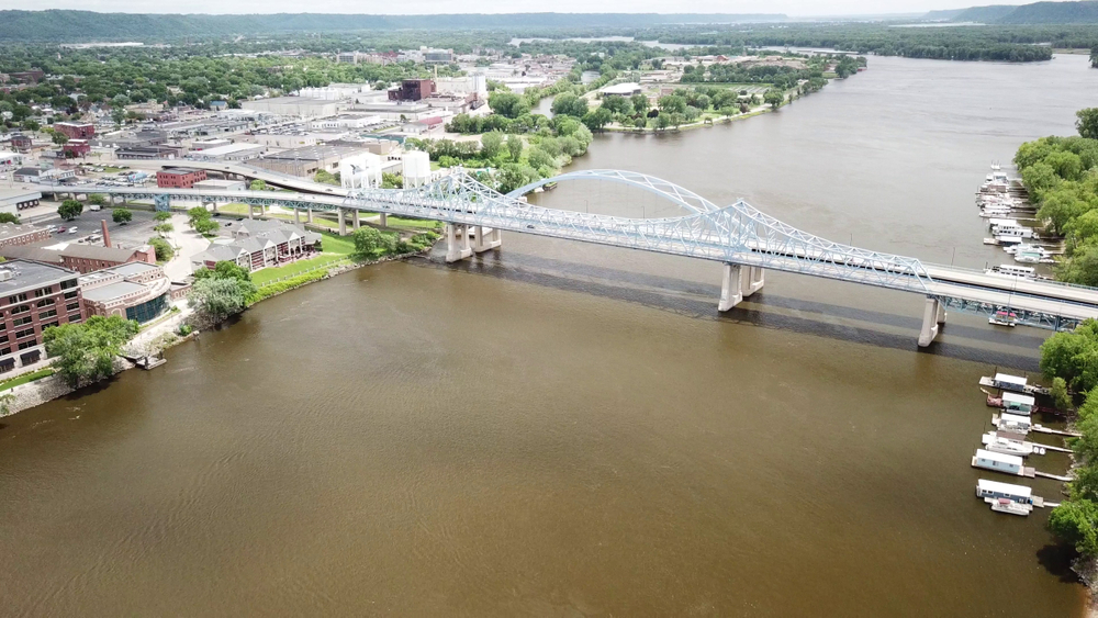 An aerial view of a large white bridge crossing the river over to LaCrosse Wisconsin. On the banks of the river you can see some house boats. On the other side of the river are lots of industrial buildings. 