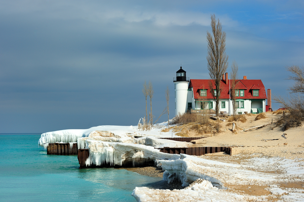A small white light and black light house with red shingles on the roof. It is on the sandy shores of a lake but there is also snow and ice hanging off the dock near the lighthouse. There are dunes with dried grass and trees with no leaves. One of the best things to do in Michigan