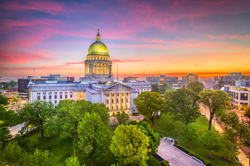 A slight aerial view of the Wisconsin Capital Building in Madison. It is a large Greek Revival building with a tall green dome in the middle. It is surrounded by a grassy park with trees. The building is lit up and the sun is setting so the sky is red, yellow, orange, pink, and purple. 