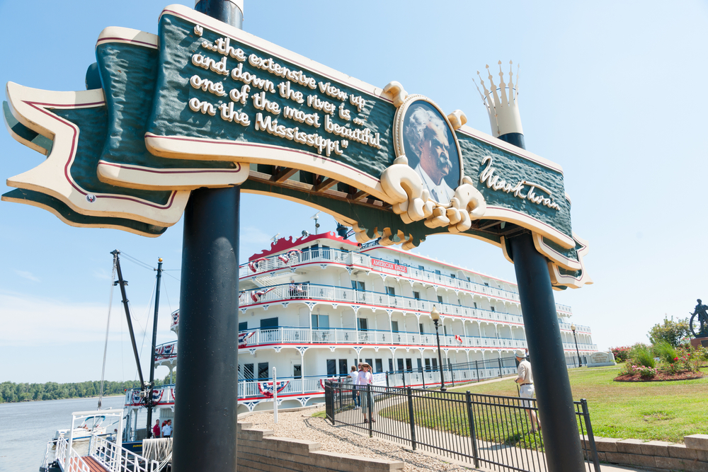 A sign with a portrait and quote from Mark Twain over a dock leading to a classic steam white and red steamboat on the Mississippi River. It is in Mark Twain's hometown. 