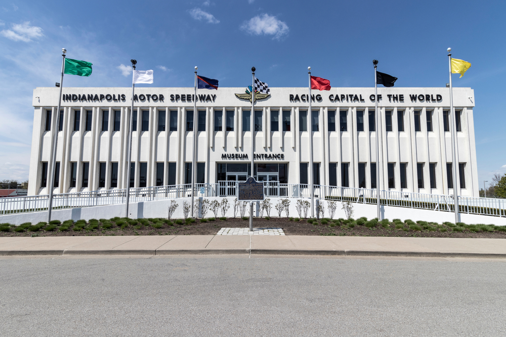The exterior of a large white builiding that is the Indianapolis Motor Speedway Museum. In front of the building is a ramp that goes up either side to the entrance. In front of the ramp are flag poles with green, white, blue, checkered, red, black, and yellow flags. 