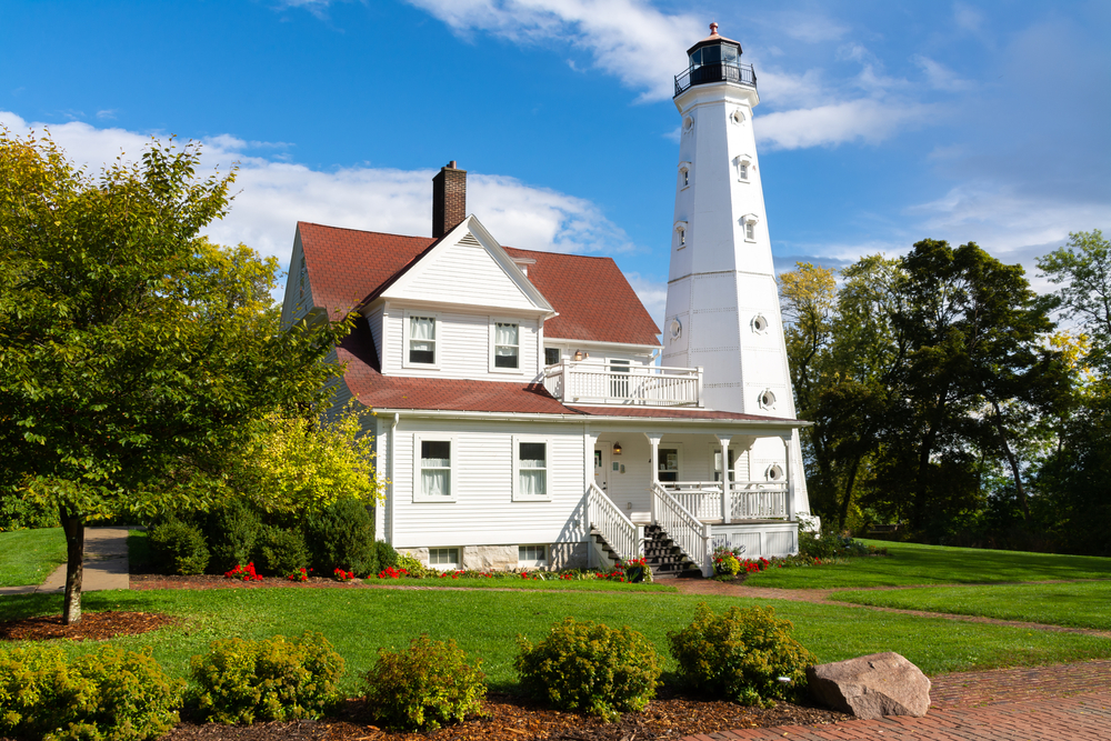 The North Point Lighthouse which is a large white house attached to a white light tower. The lighthouse has a green lawn with some red flowers, shrubs, and trees around it. One of the best things to do in Wisconsin. 