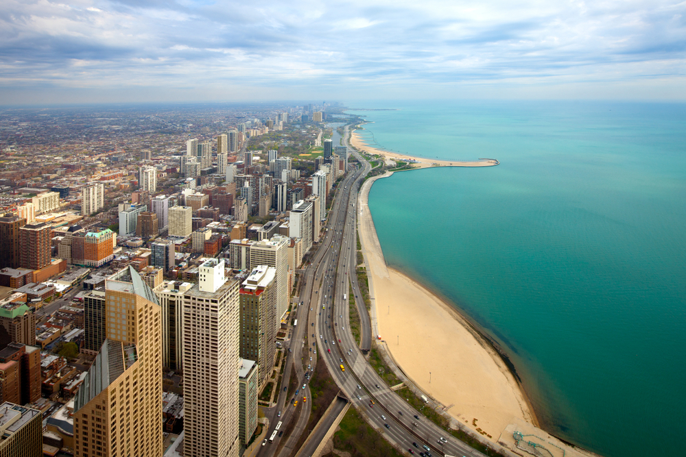 An aerial view of the Oak Street Beach in Chicago. You can see the beach on Lake Michigan and then right behind it is the city skyline of Chicago. The beach and the city extend into the horizon for miles and the water on the lake is very blue. 