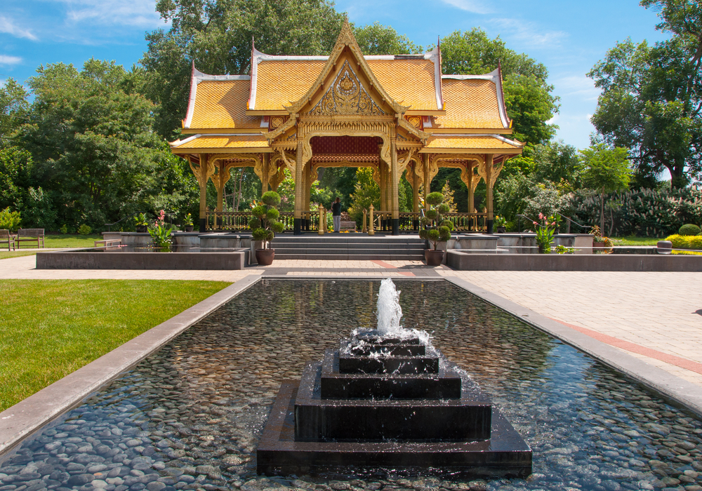 A Thai inspired pavilion in the Olbrich Botanical Gardens. The building is a creamy yellow and has two ponds next to it. There is also a bigger pond with a water feature in front of it. There are plants and a grassy area around it. 