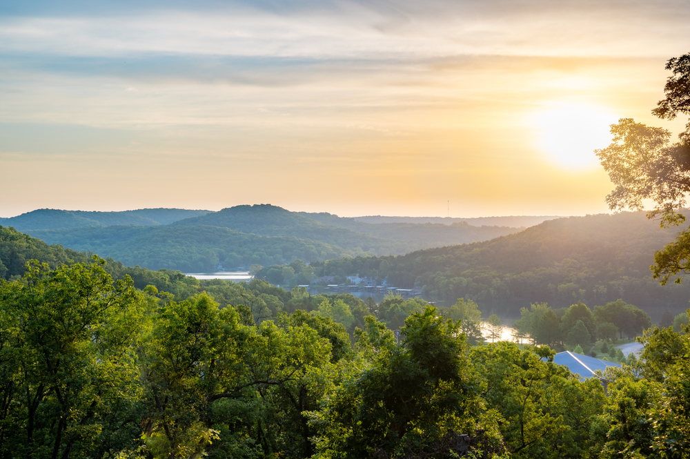 The view of the mountains in the Ozarks at sunset. The mountains are covered in green trees, you can see some water, and the roofs of some buildings. 