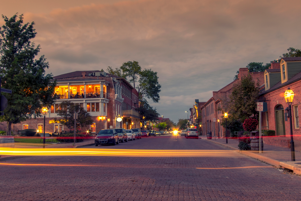 Looking down the street of St. Charles. It is twilight and the sky is dark and cloudy. The street lights are on and you can see one of the buildings lit up. There is a light trail going across the picture from a car driving by. 