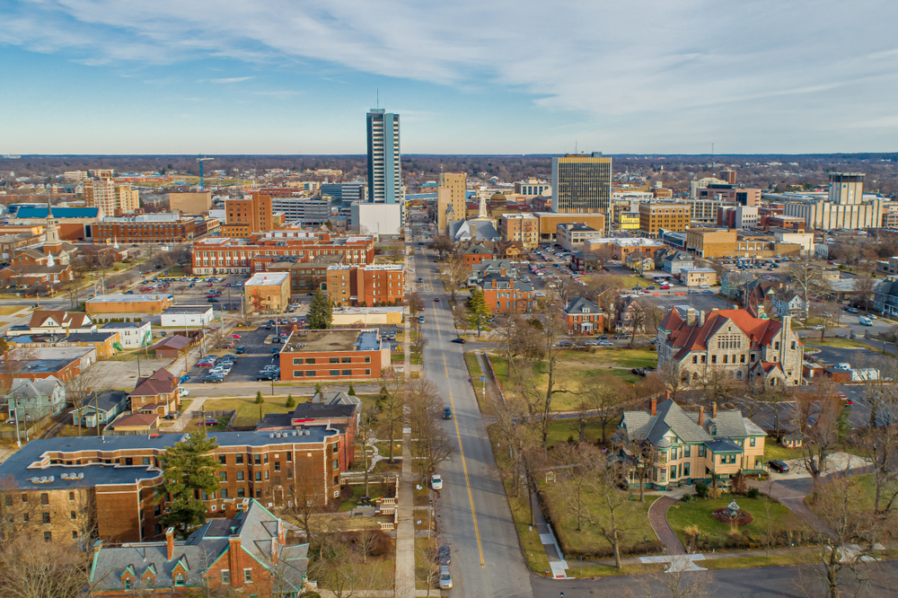 An aerial view of South Bend. There are old homes, old churches, a few small skyscrapers. You can see lawns and trees and what looks like a large forest area in the distance. 