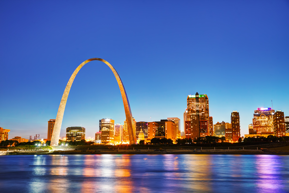 The St. Louis skyline and the Gateway Arch. It is twilight and the city is all lit up as well as the Gateway Arch. The view is from the river and the lights are reflected on the river. One of the best Missouri road trips stops.