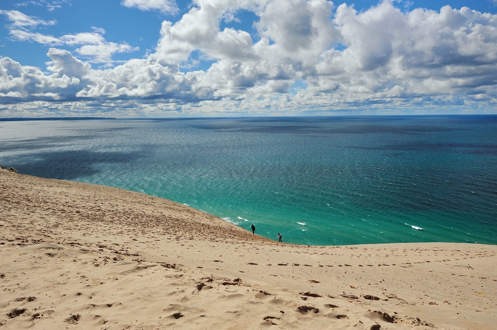 A huge sand dune leading done to crystal blue water with people walking along the shore. The Dune climb at Sleeping Bear Park one of the things to do in Traverse City