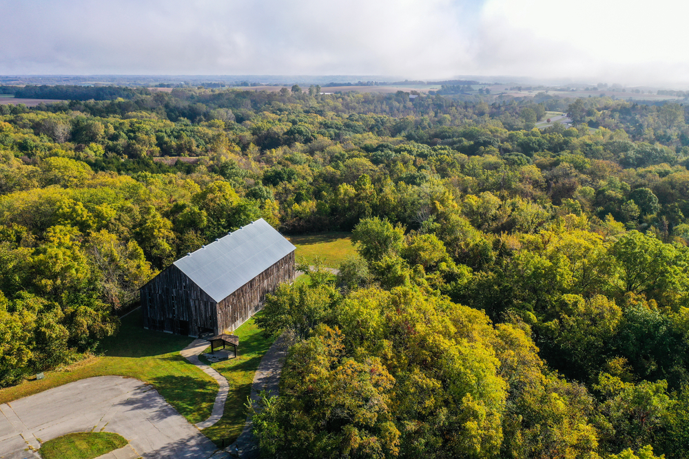 An aerial view of the Weston Bend State Park. It is covered in trees with green leaves, but in the far distance you can see fields. There is a restored barn at the front  of the image.