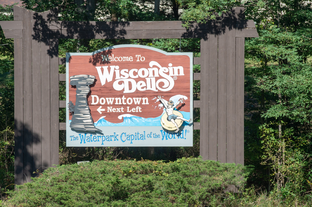 A wooden sign welcoming you to Wisconsin Dells, the Waterpark Capital of the World. There is a drawing of a rock formation and a kid on an innertube on some waves on it. 