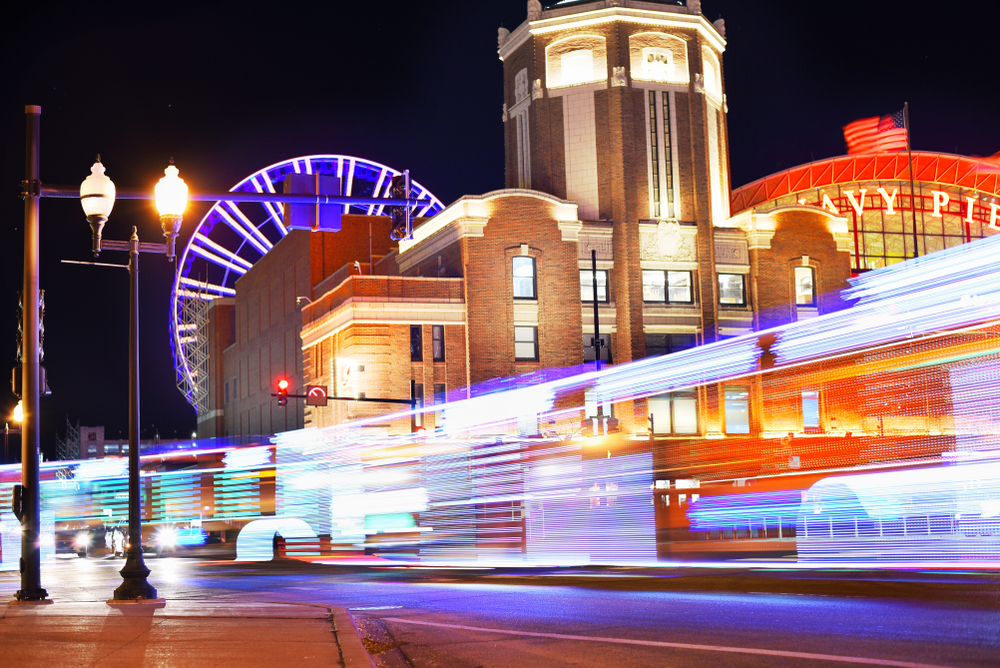 The light trail of the CTA bus in Navy Pier. The Navy Pier is all lit up and it is dark out. The area is lit up in red, blue, and yellow lights. 