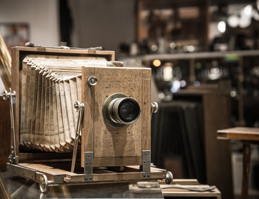 An antique accordion camera in a studio. Behind it you can see a blurry studio with benches, worktables, and what looks like other cameras. 

