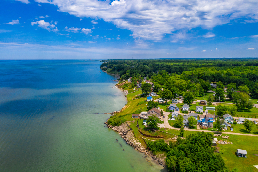 An aerial view of looking down the Lake Erie coast in Ashtabula. The water is very blue and the shoreline is mostly rocky or grassy. You can see a large area of trees and then some houses that are very close to the shore line, up a slight hill. 