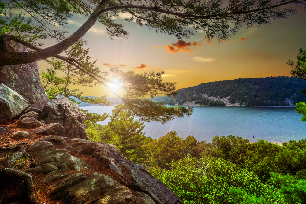 The view of Devil's Lake at Devil's Lake State Park in Baraboo. You can see a rocky cliff, trees with green leaves, a blue lake, and a hill on the other side of the lake covered in trees. 