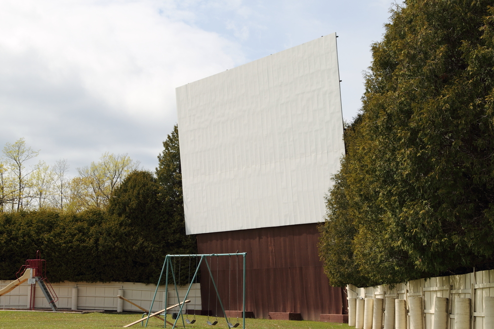 A side view of a large theater screen at an outdoor drive in movie theater. On the ground in front of the screen there is a small playground area with swings, a seesaw, and a slide. On both sides of the screen there is a row of trees. 
