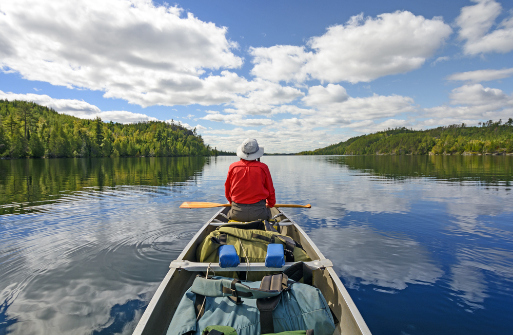 A person in a large kayak on a lake. They are wearing a red shirt and a hat. They are looking out into the distance. There are trees on either side of the lake. 