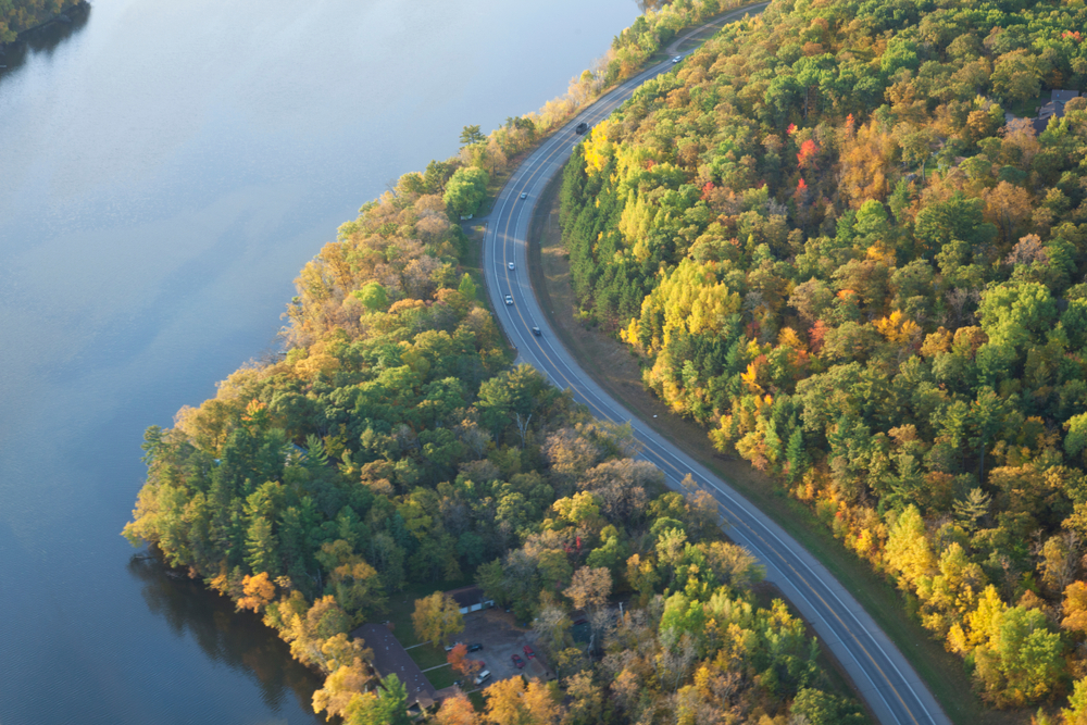 An aerial view of a twisting road near Brainerd. It is curving along the river and surrounded by trees. The trees have green, yellow, orange, and red leaves. 