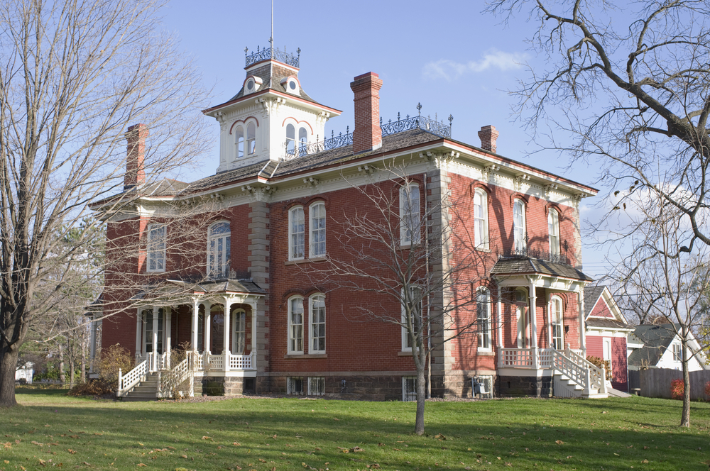 A large Victorian mansion in a Wisconsin small town. It is made of brick and has white trim. There are several staircases on it and a white section on the roof that resembles a bell tower. It has a large lawn with trees with no leaves. 