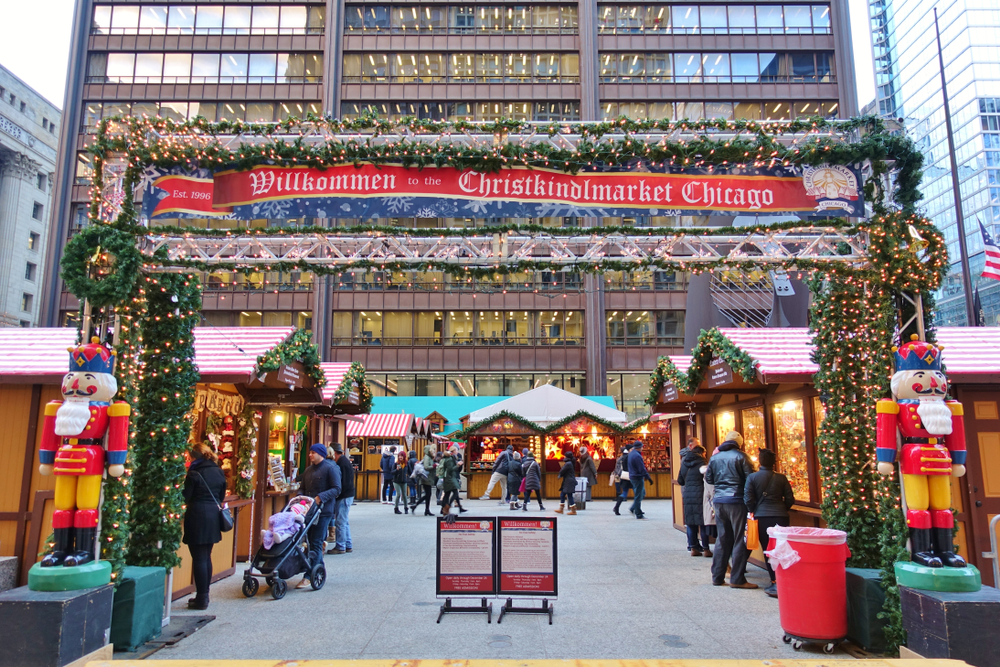 The front entrance of the Christkindlmarket in Chicago during the day. It is a large archway with a red sign on it. The arch way is covered in greenery and lights with two nutcrackers. Past the entry you can see small stalls of little Christmas hits all lit up. 