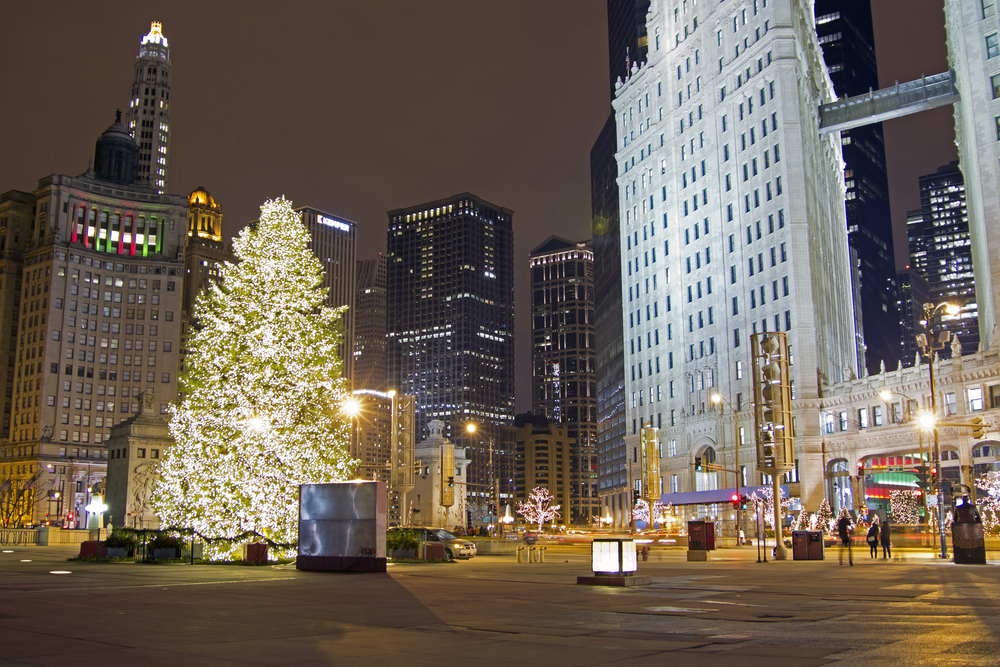 A large Christmas tree all lit up at night in Chicago's Millennium Park. There are people walking around, the shops are all lit up, and there are other trees with lights on them. 