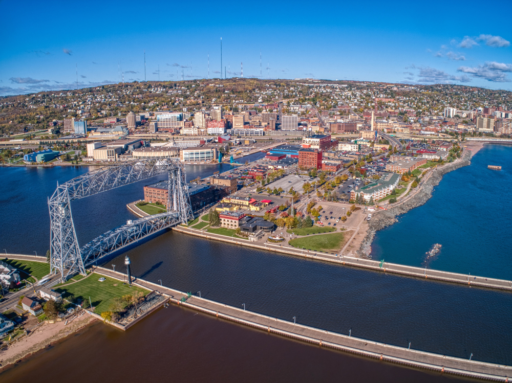 An aerial view of Duluth on the edge of the Mississippi River. You can see a large bridge, buildings along the water, and buildings in the distance. The sky is blue with scattered clouds.