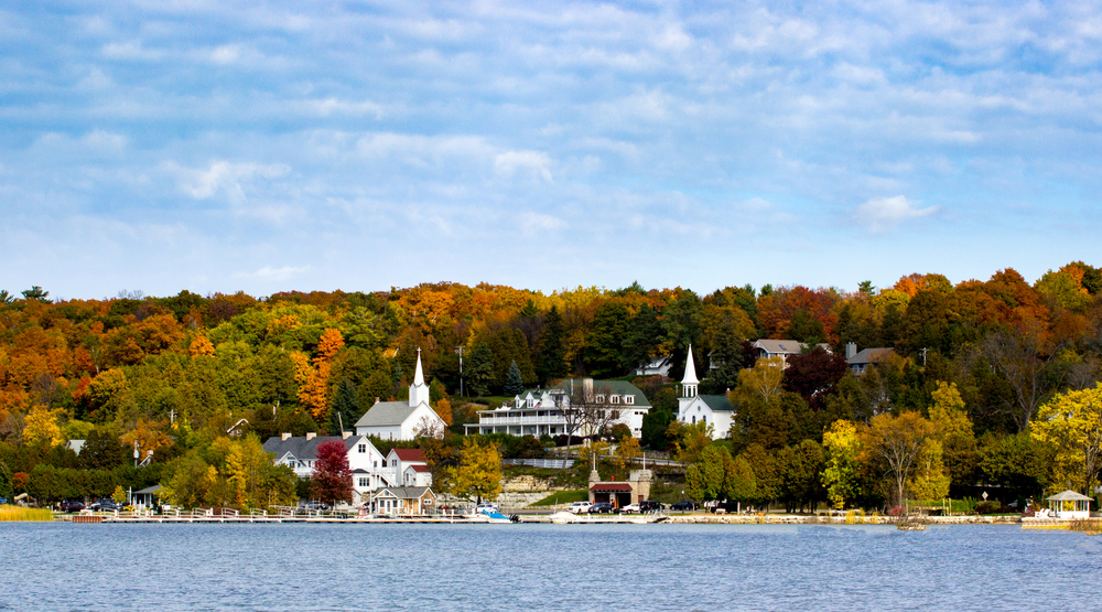 A cute Wisconsin small town being viewed from a lake. On the shore you can see a boat marina. You can also see two white churches with tall steeples, a large Victorian inn, and other buildings. The town is full of trees with red, yellow, orange, and green leaves. 