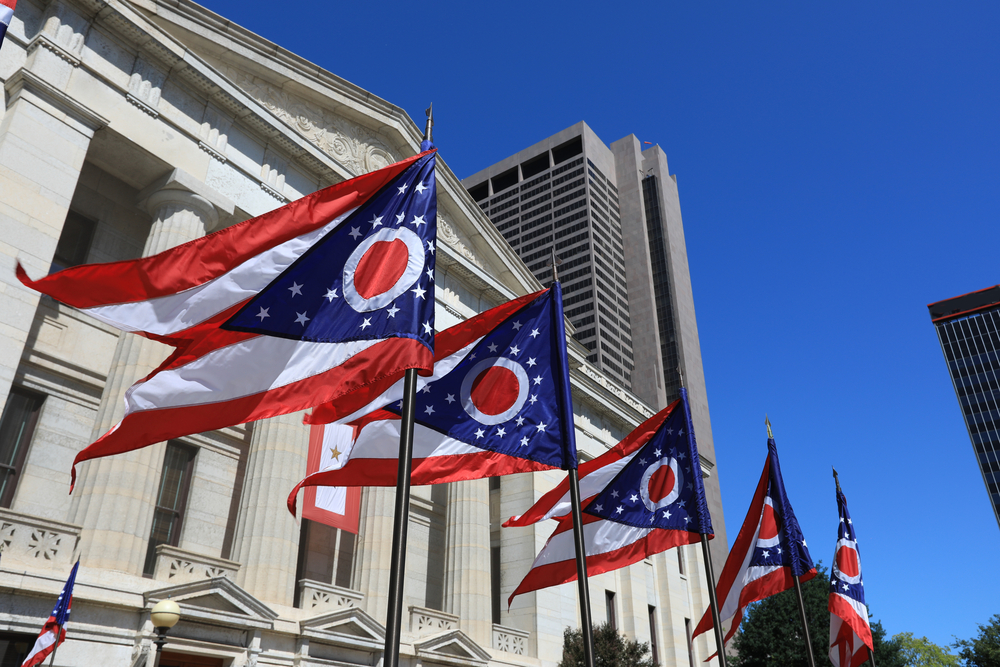 Looking up at four Ohio state flags flying in front of a government building. You can also see a few high rises. The Ohio state flag has a triangle cut out from the middle of it form the the bottom, has red and white stripes, and a blue triangle with white stars and a white O with a red center. 