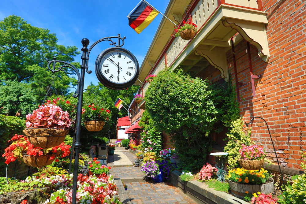 Looking down a sidewalk in the German Village of Columbus Ohio. The houses are surrounded by flower pots with flowers in several different colors. There is a clock hanging on a wrought iron shepherds hook. The buildings are brick with terraces that have German flags hanging from them. One of the best Ohio day trips.