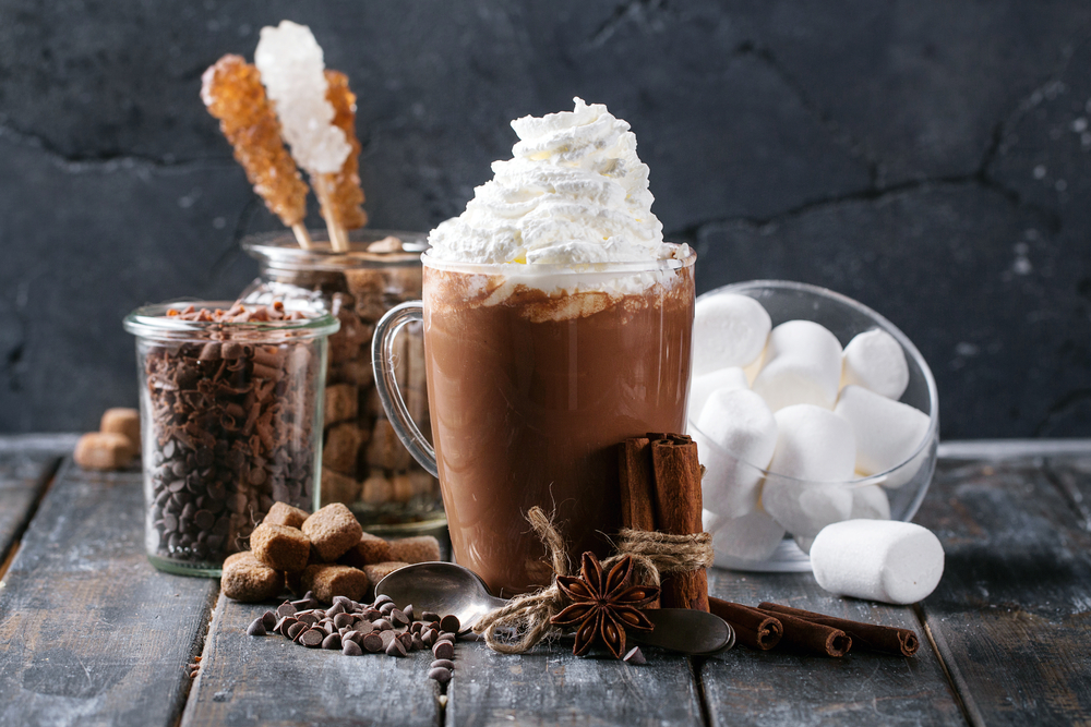 A cup of hot cocoa covered in whip cream. Behind it there are jars of marshmallows, rock candy, shaved chocolate, and other toppings. Around the clear mug there is sticks of cinnamon, anise, and a spoon. 
