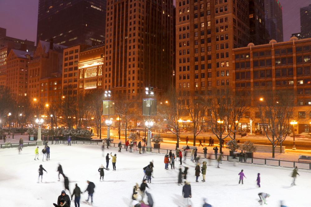 People ice skating at night in a park in Chicago. The buildings are all lit up and the street lights are on. The park is all lit up with large stadium style lights. One of the best things to do during Christmas in Chicago. 