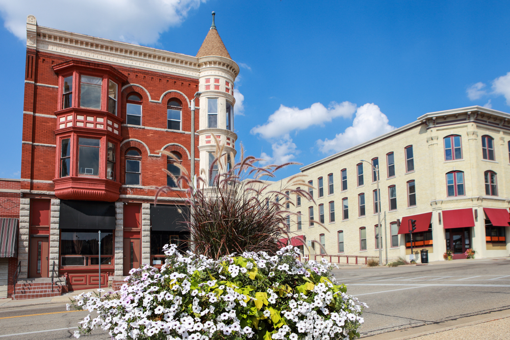 A Main Street in a small town. The buildings are all Victorian style buildings. They are made of red brick or painted a cream color. There is a large flower post with white flowers, tall fuzzy grass, and a bright green plant. 