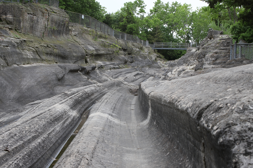 Looking down the path of the Kelleys Island Glacial Grooves. They are smoothed out rock grooves in a dark grey rock. The grooves all fluctuate in depth and shape. In the distance you can see a metal bridge with trees behind it crossing the grooves. One of the best Ohio day trips.