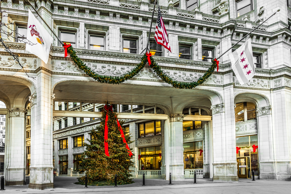 A large ornate shopping district entrance of the Magnificent Mile in Chicago. In the middle of the ornate arch way entrance is a large Christmas tree lit up with a big red bow on it. There is also a sway of greenery with red bows and lights hanging on the archway. 