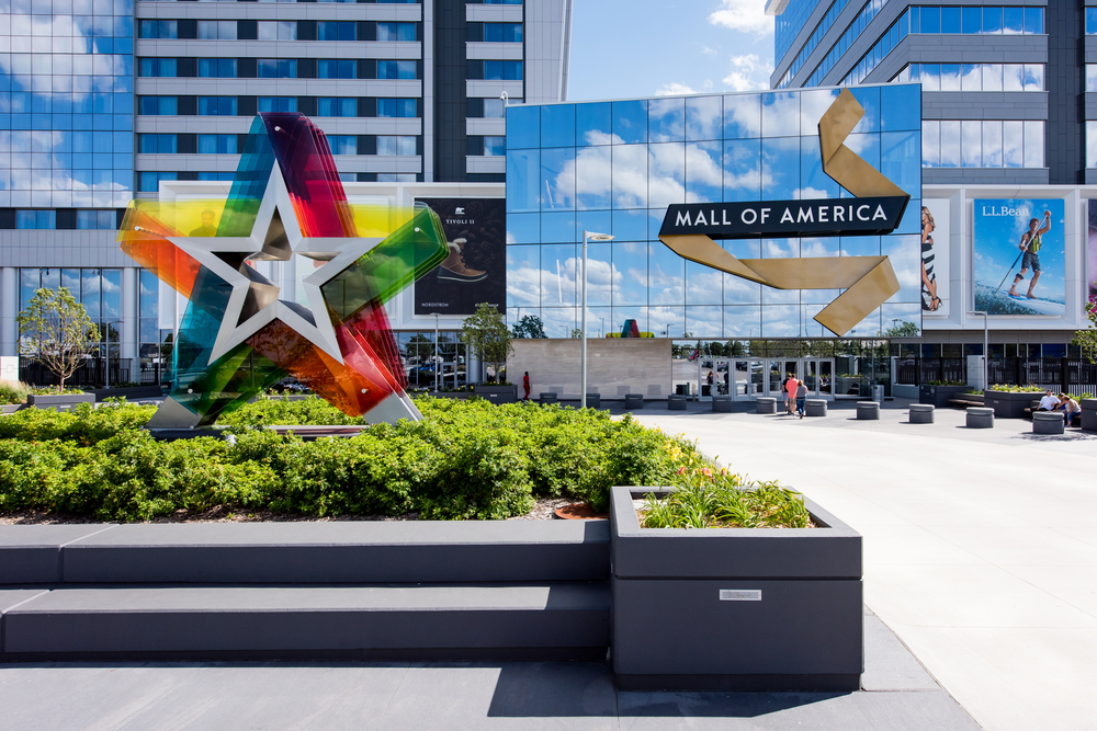 The exterior entrance to the Mall of America. IT is a large multi level building with windows that are reflecting the sky. In a area with small shrubs there is a large rainbow colored translucent star. One of the best things to do in Minnesota.