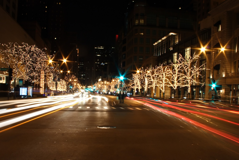 A main street in Chicago in the winter. There are trees lining the street and they are covered in twinkle lights that are all lit up. It is night time and on the street you can see light streaks from cars passing by. 