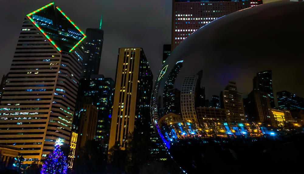 The famous bean in Millennium Park. It is dark out and the city is all lit up with holiday lights. The skyline and the lights are reflected on the surface of the bean sculpture. 